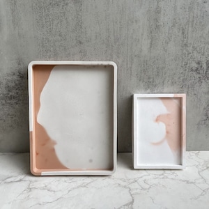 May include: Two rectangular trays made of concrete with a white and light pink marbled design. The larger tray is on the left and the smaller tray is on the right.
