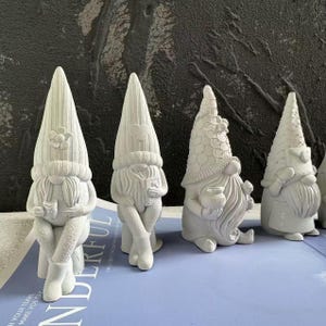 Könnte beinhalten: Vier weiße Gartenzwerge mit spitzen Hüten und langen Bärten. Jeder Zwerg hat ein einzigartiges Design, darunter eine Blume, ein Herz und strukturierte Muster. Ein Zwerg sitzt und hält eine Tasse. Die Zwerge stehen auf einer blauen Oberfläche.