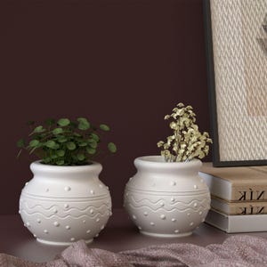 Puede incluir: Dos jarrones de cerámica blancos con un patrón ondulado y de puntos. Uno contiene plantas verdes con hojas, el otro flores doradas secas. Los jarrones están sobre una superficie con una tela morada, con libros y un cuadro en el fondo.