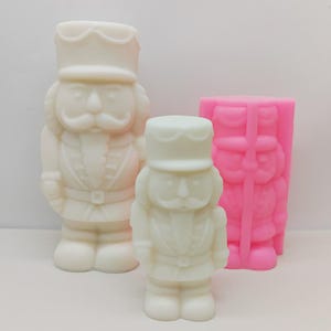 Könnte beinhalten: Drei Nussknacker-Figuren in verschiedenen Größen. Zwei sind cremefarben und einer ist rosa. Die Nussknacker haben detaillierte Merkmale, darunter Hüte, Schnurrbärte und Jacken im Militärstil. Der rosafarbene scheint eine Form zu sein.