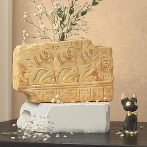 May include: A beige rectangular vase with ancient Egyptian carvings, holding white flowers. It rests on a white block, with a black cat figurine with gold accents nearby. The vase features hieroglyphs and figures.
