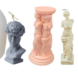 May include: Three decorative candles shaped like classical sculptures. The first candle is a bust of a man with a laurel wreath on his head. The second candle is a column with two women standing back to back. The third candle is a statue of Venus, a woman standing with a drapery. The Venus candle has the word "VENUS" written on the base.