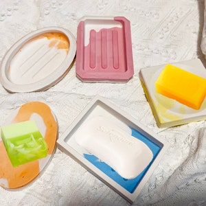 Könnte beinhalten: Eine Sammlung farbenfroher Seifenschalen in verschiedenen Formen und Designs. Die Schalen sind aus Beton gefertigt und enthalten Seifenstücke in Grün-, Gelb- und Weißtönen. Die Seifenschalen sind oval, rechteckig und quadratisch.