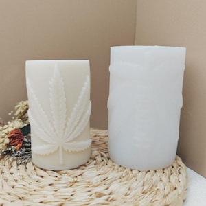 Könnte beinhalten: Zwei weiße Säulenkerzen. Eine Kerze hat ein detailliertes Cannabisblatt-Design. Die andere ist eine leere Form. Die Kerzen stehen auf einer gewebten Matte mit getrockneten Blumen. Der Hintergrund ist ein neutrales Beige.