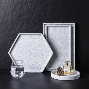 May include: Three white speckled ceramic trays, one hexagonal, one rectangular, and one round. The round tray has a small pine cone and a bottle of perfume on it.