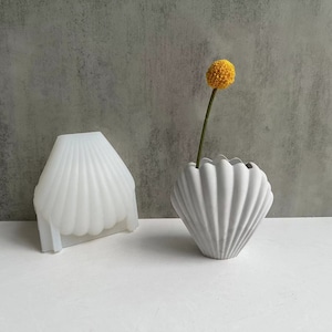 May include: Two white ceramic seashell vases, one with a yellow flower, on a white surface.