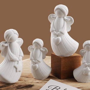 Könnte beinhalten: Vier weiße Keramik-Engelsfiguren, jede mit detaillierten Flügeln und fließenden Gewändern. Zwei Engel halten Herzen und zwei Sterne. Ein Engel wird auf einem kleinen Holzblock ausgestellt. Die Engel sind auf einer Holzoberfläche angeordnet.