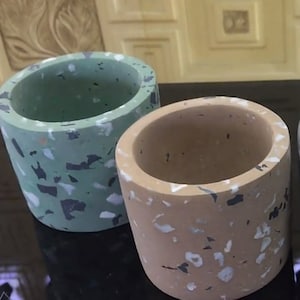 以下が含まれることがあります： 斑点のあるテラゾーデザインの小さなコンクリートプランター2つ。1つは緑色、もう1つはベージュです。