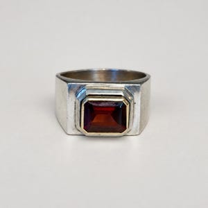 May include: A silver ring featuring a rectangular, deep red garnet gemstone set in a gold-toned bezel. The band is wide and smooth, with a polished metallic finish that contrasts with the dark, faceted stone centered on the front of the ring.