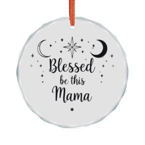 May include: A round, white ornament with the text "Blessed be this Mama" in black script. The design includes a crescent moon, stars, and a starburst. It hangs from a red ribbon.