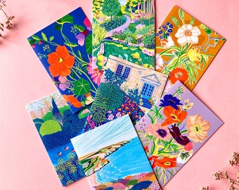Op de afbeelding: Een verzameling kleurrijke notitieboekjes met bloemen- en landschapsillustraties. De notitieboekjes hebben levendige ontwerpen van tuinen, kustlandschappen en bloemen in tinten blauw, oranje, geel en paars. De notitieboekjes zijn gerangschikt op een roze ondergrond.