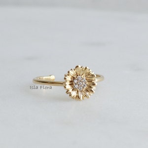 May include: A gold ring featuring a small, round, clear stone set in the centre, surrounded by a textured, flower-shaped design.