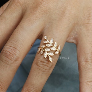 May include: A gold ring with a leaf design featuring small clear stones. The ring is on a finger with light pink nail polish.