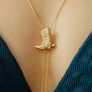 May include: A gold-toned lariat necklace featuring a detailed cowboy boot pendant. The delicate chain threads through the boot, ending in two dangling strands with small spherical beads at the tips, resting against a dark teal textured fabric.
