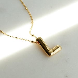 May include: A gold-coloured necklace featuring the letter "L" pendant. The chain is delicate with small, evenly spaced beads. The pendant is a solid, stylised letter "L". The necklace is displayed on a white surface with bright light.