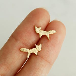 May include: A pair of small, gold-colored fox-shaped stud earrings. The earrings are held on a person's finger. The fox design is simple, with a textured surface and a small tail. The earrings are likely made of metal.