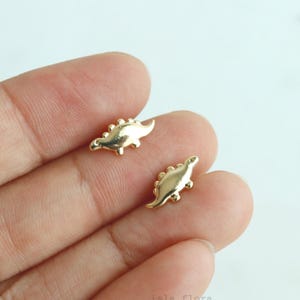 May include: Two small, gold-colored dinosaur charms. Each charm has a detailed, textured design, including a row of small spikes along the back. The charms are displayed against a light background.