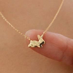May include: A gold-toned necklace featuring a small, polished rabbit-shaped pendant suspended from a delicate chain. The pendant is smooth and reflective, capturing light on its curved surface, and is attached to the chain at the top of the rabbit's ears.