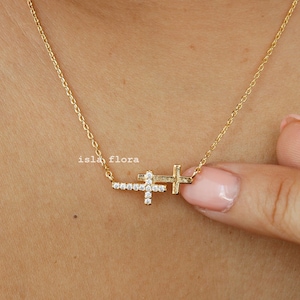 May include: A gold chain necklace with two small, diamond-encrusted crosses. The crosses are layered on top of each other, with the top cross slightly offset to the right. The necklace is worn by a person with fair skin.