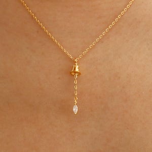 May include: A delicate gold-colored necklace featuring a small bell pendant with a dangling, teardrop-shaped clear crystal. The necklace is displayed against a skin-toned background, highlighting the jewelry's design.