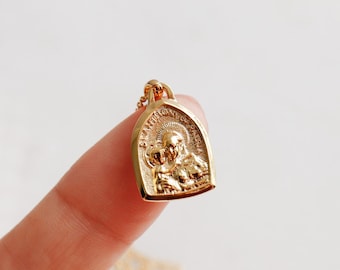 May include: A small gold-toned pendant featuring an embossed religious scene of two figures with halos, shaped like an arch with a rounded top. The pendant is attached to a delicate gold-toned chain, showcasing intricate metalwork and a polished finish.