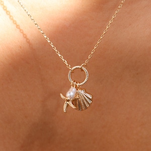 May include: A gold-toned chain necklace featuring a circular pendant charm. The charm is adorned with a small white pearl, a textured gold starfish, and a gold seashell. The necklace rests against skin, showcasing the delicate metal link design.