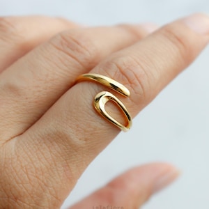 May include: A gold-toned open ring featuring a smooth, polished finish. The design consists of a curved band that wraps around the finger, ending in a rounded, teardrop-shaped loop that remains open, creating a continuous, fluid metal silhouette.
