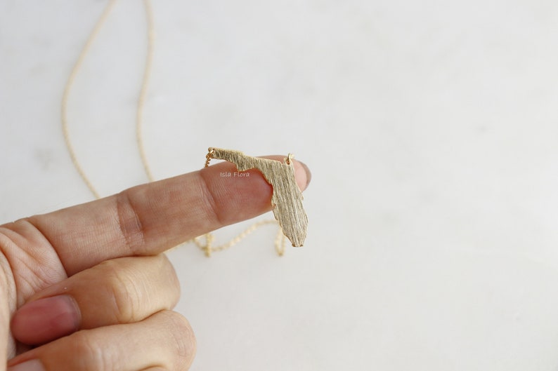 May include: Gold necklace featuring a textured outline of the state of Florida. The necklace is on a white background.
