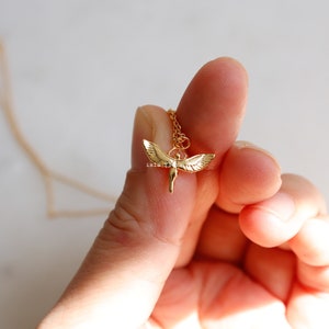 May include: A gold-toned necklace featuring a small pendant shaped like a figure with outstretched wings. The pendant is suspended from a delicate gold-colored chain. The metal has a polished finish that reflects light, highlighting the wing details.