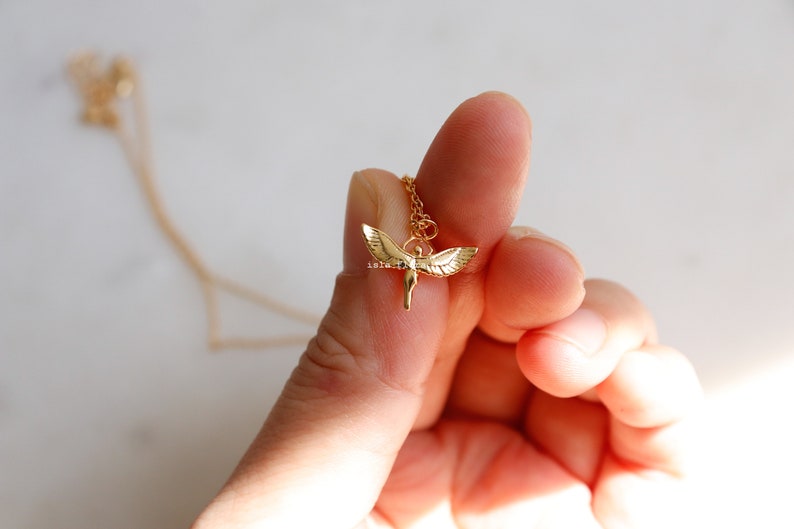 May include: A gold-toned necklace featuring a small pendant shaped like a figure with outstretched wings. The pendant is suspended from a delicate gold-colored chain. The metal has a polished finish that reflects light, highlighting the wing details.