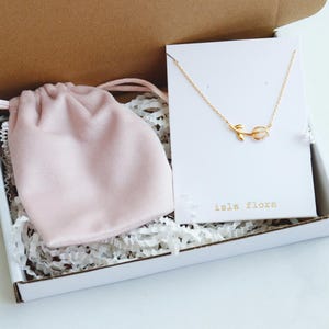 May include: A gold-toned necklace featuring a small tulip pendant with a white stone, displayed on a white card labeled "isla flora" inside a cardboard box. A soft pink drawstring pouch rests next to the necklace on white paper filler.