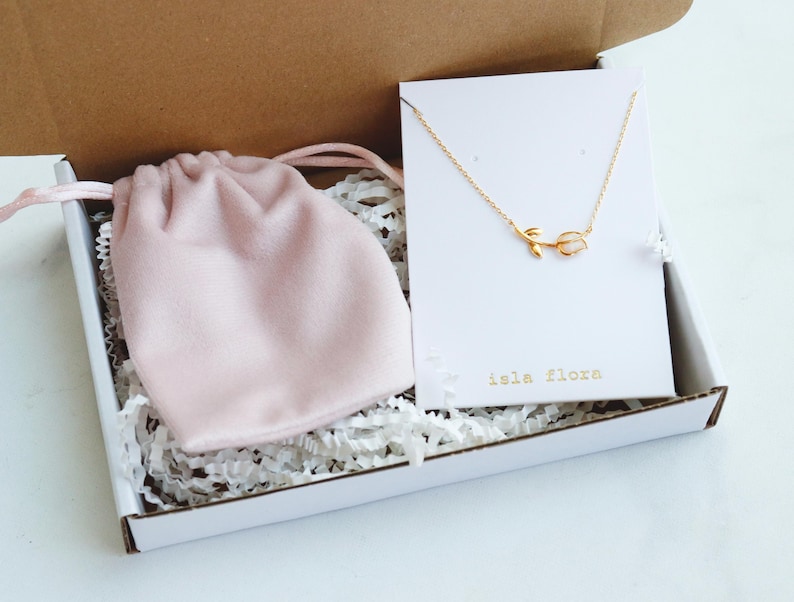 May include: A gold-toned necklace featuring a small tulip pendant with a white stone, displayed on a white card labeled "isla flora" inside a cardboard box. A soft pink drawstring pouch rests next to the necklace on white paper filler.