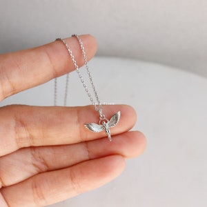 May include: A silver-toned necklace featuring a delicate chain and a small pendant shaped like a figure with outstretched wings. The metal has a polished finish, and the pendant is suspended from a simple jump ring attached to the thin link chain.