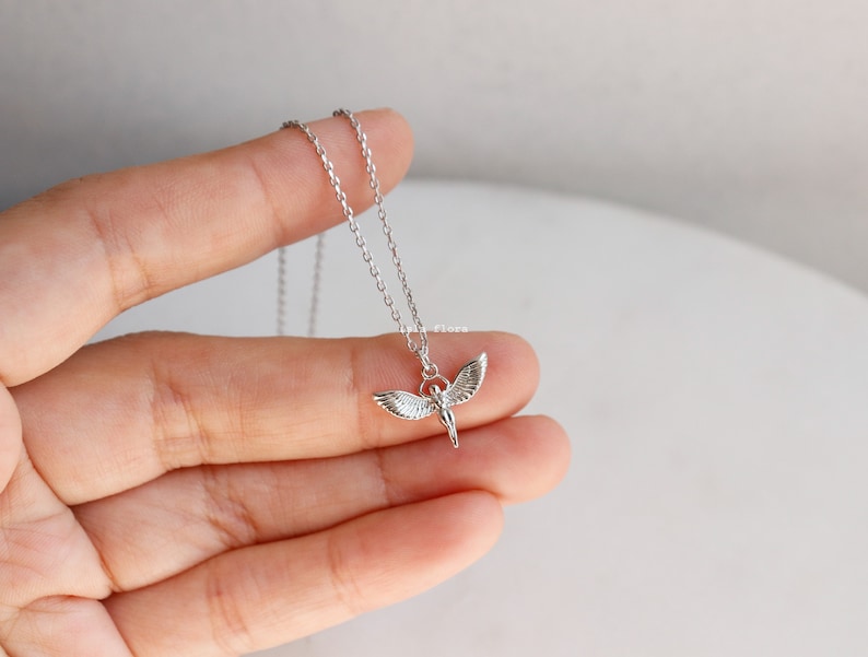 May include: A silver-toned necklace featuring a delicate chain and a small pendant shaped like a figure with outstretched wings. The metal has a polished finish, and the pendant is suspended from a simple jump ring attached to the thin link chain.