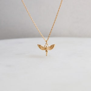 May include: A gold-toned necklace featuring a delicate chain and a small pendant shaped like a figure with outstretched wings. The pendant is suspended from the center of the chain, showcasing detailed feather textures on the wings and a smooth body.