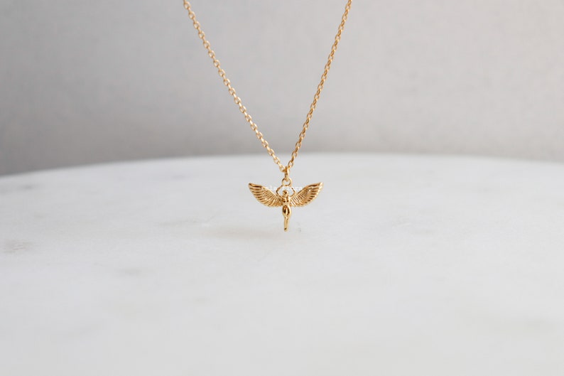May include: A gold-toned necklace featuring a delicate chain and a small pendant shaped like a figure with outstretched wings. The pendant is suspended from the center of the chain, showcasing detailed feather textures on the wings and a smooth body.
