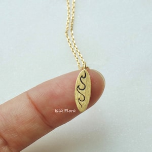 Puede incluir: Collar con colgante de tabla de surf dorado. El colgante presenta una superficie texturizada con un diseño de olas negras. El collar está en una delicada cadena dorada. La marca "Isla Flora" es visible.