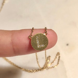 May include: A gold-colored necklace featuring a circular pendant with the letter "L" encrusted with small, sparkling stones. The pendant hangs from a delicate chain. The necklace is shown against a neutral background.