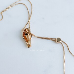 May include: A gold-toned necklace featuring a horse head pendant with a hollow center. The chain is a delicate box link style with an adjustable sliding bead mechanism. The text "isla flora" is printed in small white letters below the pendant.