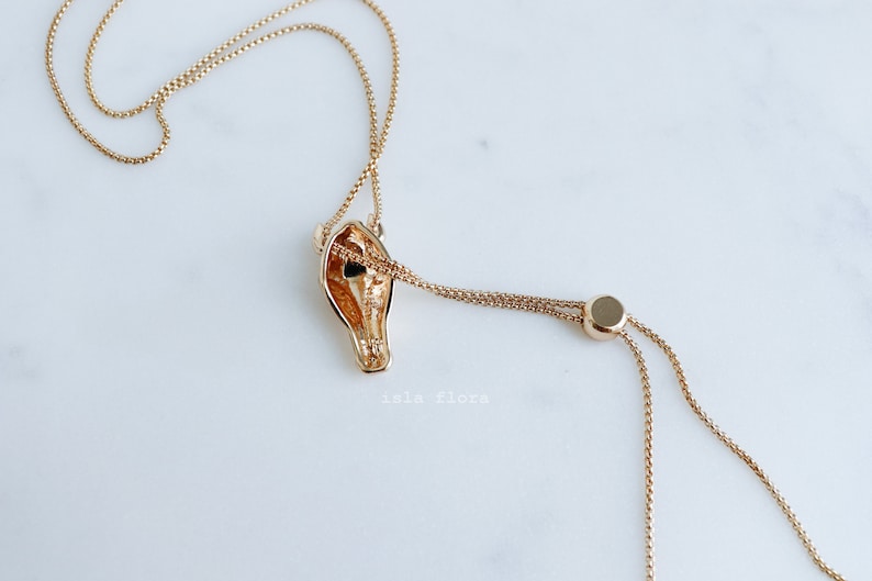 May include: A gold-toned necklace featuring a horse head pendant with a hollow center. The chain is a delicate box link style with an adjustable sliding bead mechanism. The text "isla flora" is printed in small white letters below the pendant.