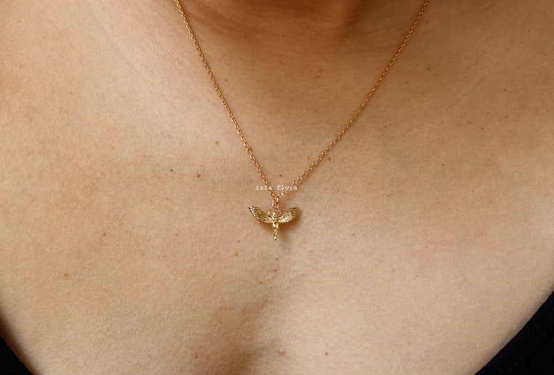 May include: A gold-toned necklace featuring a delicate chain and a small pendant shaped like a winged figure with outstretched wings. The pendant is centered on the chain, which rests against the skin. The text "isla flora" is visible above the pendant.