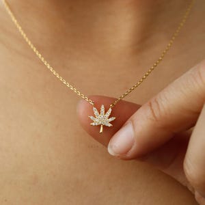 May include: A gold-toned necklace featuring a delicate chain and a small pendant shaped like a cannabis leaf. The leaf pendant is encrusted with numerous tiny, clear, sparkling stones that cover its entire surface, creating a bright, reflective appearance.