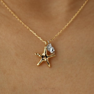 May include: A gold-toned necklace featuring a delicate chain and a pendant shaped like a five-pointed starfish. Attached to the starfish is a small, clear, faceted crystal charm that catches the light. The metal has a polished, reflective finish.