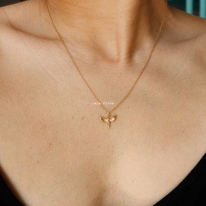 May include: A gold-toned necklace featuring a delicate chain and a small, detailed pendant shaped like a moth with outstretched wings. The pendant is centered on the chain, which rests against the collarbone. The text "isla flora" is visible.