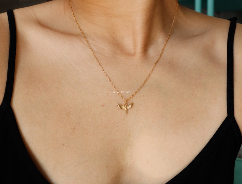 May include: A gold-toned necklace featuring a delicate chain and a small, detailed pendant shaped like a moth with outstretched wings. The pendant is centered on the chain, which rests against the collarbone. The text "isla flora" is visible.