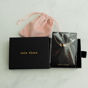 Op de afbeelding: Een zwarte doos met de tekst "isla flora" op de voorkant. De doos is open en bevat een kleine, delicate gouden ketting met een roosknop-hanger. De ketting zit in een doorzichtige plastic zak. De doos staat op een wit oppervlak.
