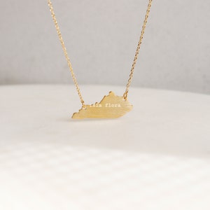 May include: A gold necklace with a pendant shaped like the state of Virginia. The pendant has the words "isla flora" inscribed on it.