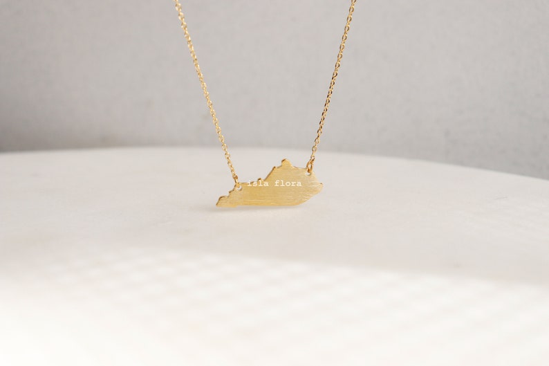 May include: A gold necklace with a pendant shaped like the state of Virginia. The pendant has the words "isla flora" inscribed on it.