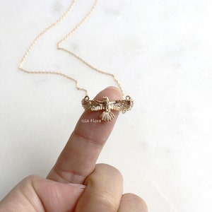 May include: A gold-coloured necklace with an eagle pendant, wings outstretched, on a fine chain. The necklace is held by a finger against a white marble surface. The brand name "isla flora" is visible. The eagle pendant is a detailed design.