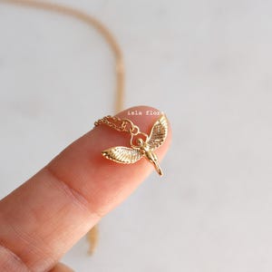 May include: A gold-toned necklace featuring a small pendant shaped like a winged figure with outstretched wings. The pendant is attached to a delicate gold chain. The text "isla flora" is printed in small, light-colored letters above the pendant.